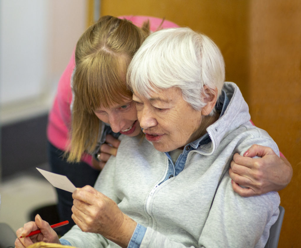 A woman hugging an elderly woman from behind. They are reading a ticket that the elderly woman is holding.